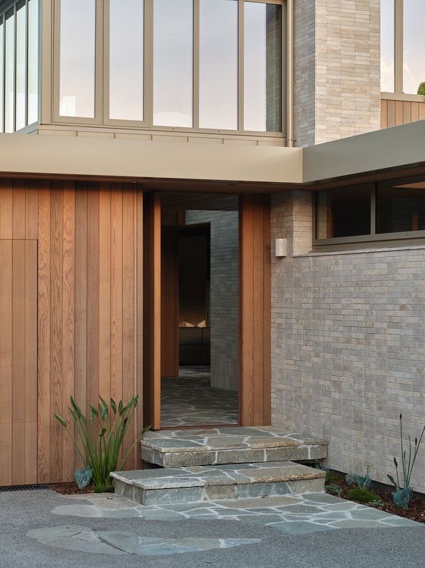 A close exterior view of a contemporary residential entry. The facade combines vertical timber cladding, pale grey brick masonry, expansive upper-level windows in slim dark frames, and broad projecting concrete or rendered forms. Irregular natural stone paving forms the path and steps leading to a timber-framed entry door, with restrained planting at the edges.