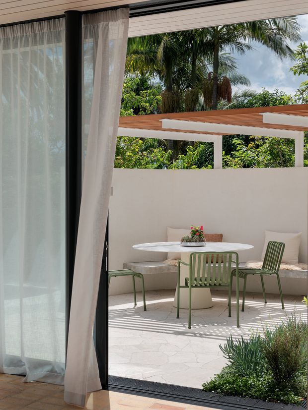 A bright indoor-outdoor view through a large black-framed glazed opening with sheer white curtains, looking onto a sheltered courtyard patio. The patio has a built-in cream bench with cushions, a round white table, and green slatted metal chairs. A low cream wall, timber-toned overhead pergola or roof element, patterned pale paving, and lush tropical planting create a relaxed resort-like setting.