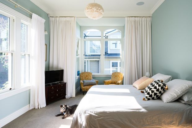 Bright, spacious bedroom with soft sage-green walls, white trim, sheer white curtains, a large bed, and two patterned armchairs by the windows. The room includes a dark timber TV cabinet, a decorative pendant light, and abundant natural light.