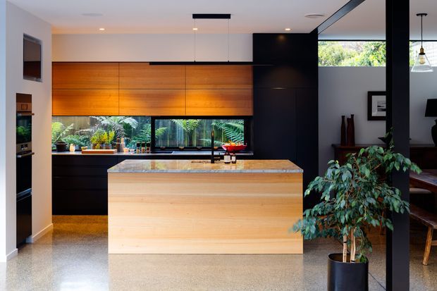 A contemporary open-plan kitchen centered on a large light timber island with a stone benchtop. The kitchen features warm horizontal timber cabinetry, dark cabinetry and structural elements, integrated appliances, a broad window with indoor plants, polished concrete-style flooring, and a dining area visible beyond a dark framed opening.