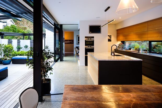 Contemporary open-plan kitchen and dining area with a large timber dining table, dark cabinetry, a pale stone island with integrated sink, and extensive glazing opening to a landscaped outdoor area. Warm timber joinery contrasts with black-framed windows and dark finishes.