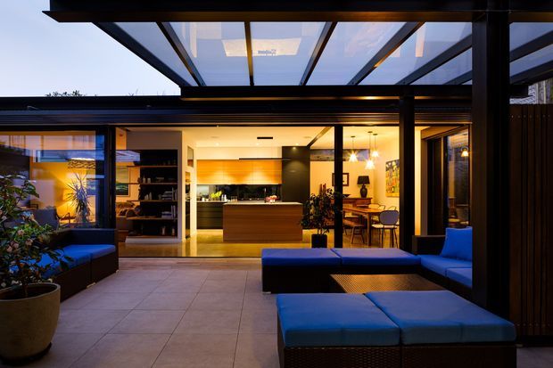 A dusk view from a covered outdoor terrace into a contemporary open-plan home. The space features a large glazed opening, dark metal framing, a transparent roof canopy, tiled terrace flooring, blue outdoor lounge seating, and a warm-lit kitchen and dining area beyond.