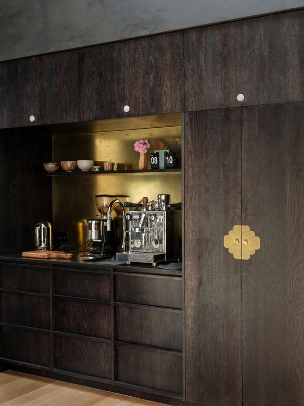A dark, richly detailed built-in coffee station concealed within full-height timber cabinetry. The cabinetry has deep brown woodgrain fronts with small round knobs and a decorative brass clover-shaped pull on the tall door. An open niche is illuminated with warm lighting and contains an espresso machine, grinder, kettle, cups and decorative objects. A small portion of light timber flooring is visible at the bottom.