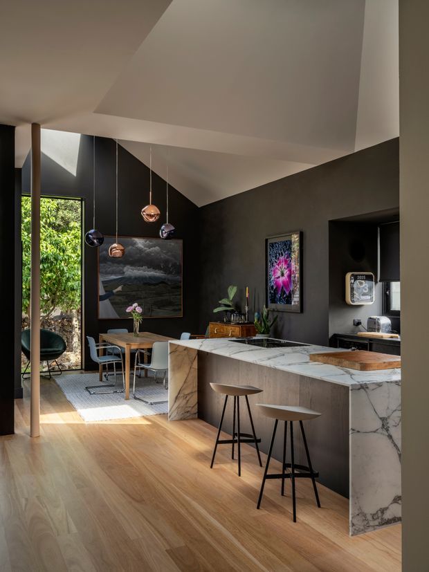 A sophisticated open-plan interior combining a kitchen and dining area. The space features warm timber flooring, dark charcoal walls, a large white-and-grey veined marble kitchen island with waterfall ends, dark cabinetry, timber dining furniture, sculptural pendant lights, and floor-to-ceiling glazing opening toward greenery. A compact coffee or service nook is visible at the rear right. The high, angular ceiling and strong natural light create a dramatic yet refined atmosphere.