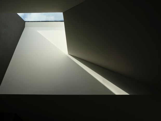 Abstract architectural interior view of a stark, sculptural lightwell or stair void. A rectangular skylight illuminates smooth pale walls and creates sharply angled geometric beams of light, contrasted by deep charcoal shadowed surfaces. No furnishings are visible.