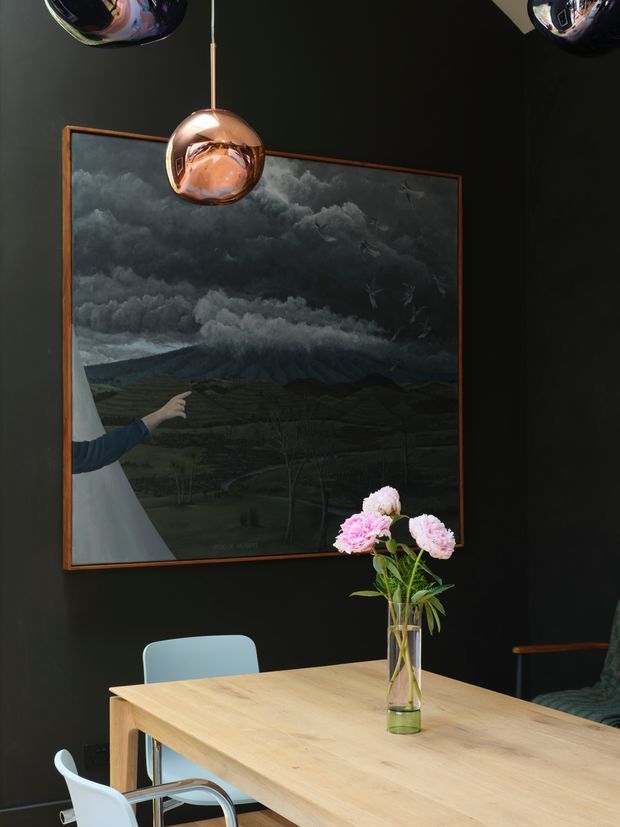 A moody dining area with a light timber dining table, pale blue and white chairs, and a clear vase of pink flowers. The room has near-black walls, a large dark landscape artwork in a timber frame, and sculptural copper pendant lights. The overall composition is dramatic and refined, with a strong contrast between the dark backdrop, warm timber, and reflective metallic lighting.