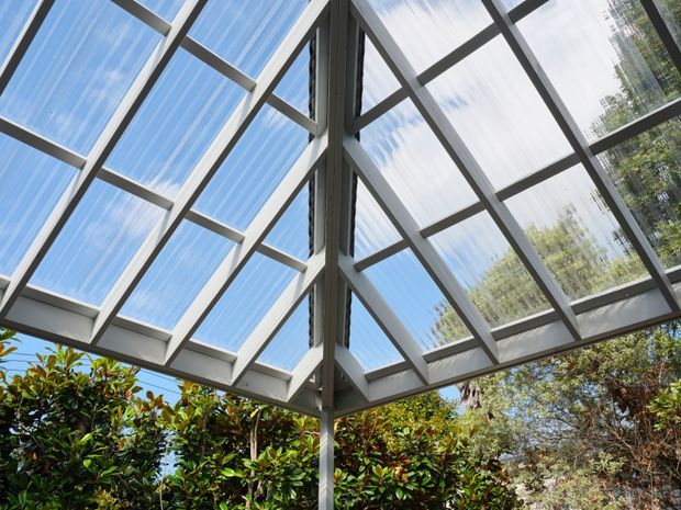 Upward-looking view of a glazed roof or conservatory-like structure with a geometric grid of grey metal framing and translucent roof panels. Blue sky and mature greenery are visible through the glazing, creating a bright, open architectural canopy.