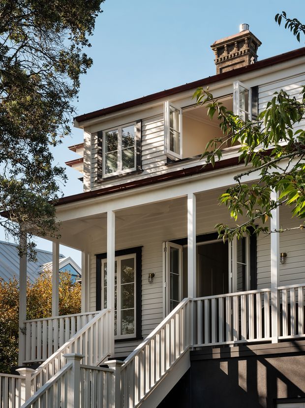 Exterior view of a two-storey light-painted weatherboard house with a deep covered front porch, white railings, open upper-level windows, dark trim and a prominent masonry chimney. Mature trees partially frame the facade.