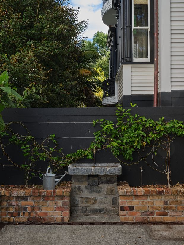 A close exterior view of a heritage-style residential building beside a landscaped courtyard or garden. The facade features dark painted weatherboard cladding, white trim, black detailing, a projecting curved balcony with decorative balustrade, and tall timber-framed windows. In the foreground is a low black-painted wall, an aged red brick retaining wall, climbing vines, a small stone-and-concrete garden water feature, and a metal watering can.