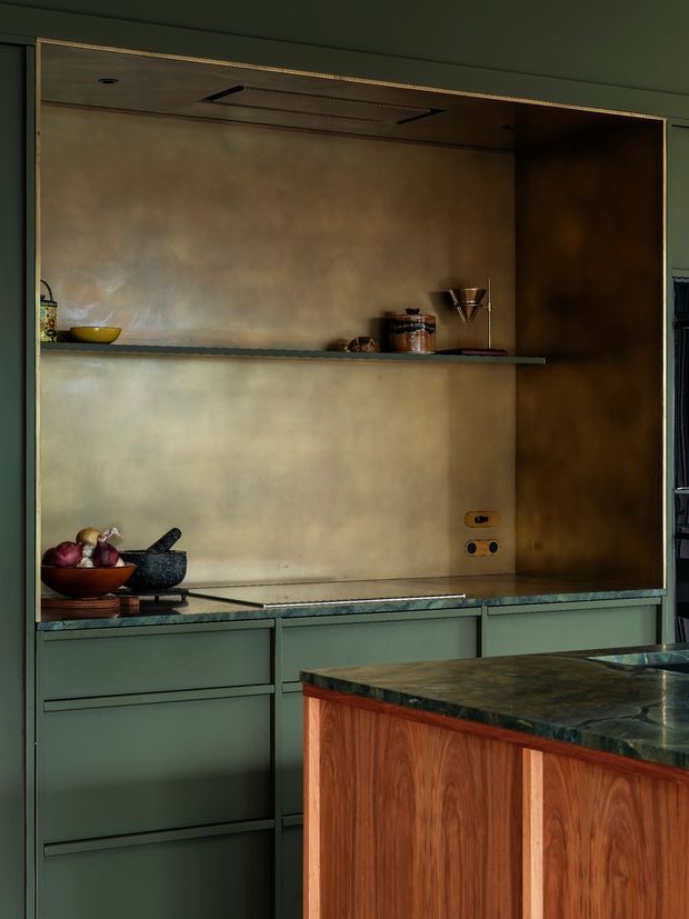 Moody close-up of a refined contemporary kitchen, featuring muted green built-in cabinetry, a dark stone worktop and island, warm timber island paneling, and a large aged brass or bronze metallic backsplash with integrated open shelving and ventilation. The composition highlights layered cabinetry, warm material contrast, and understated decorative objects.