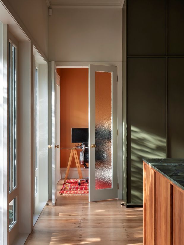A warm, sunlit interior hallway with wide timber flooring, tall glazed doors and windows on the left, a richly dark green built-in wall on the right, and a wood-fronted stone-topped counter in the foreground. A frosted-glass door opens to a small orange-red room containing a television and a patterned rug. The space features traditional architectural detailing around the ceiling and doors, with strong contrast between pale timber, deep green joinery, and terracotta accents.