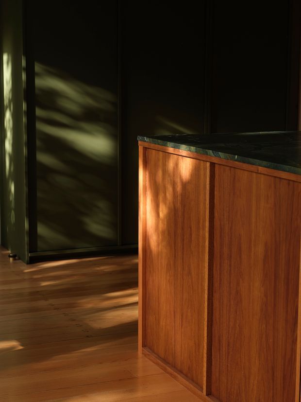 Moody close-up of a timber kitchen island or built-in counter with richly grained warm wood panelled cabinetry and a dark stone benchtop. The space has dark wall cabinetry or panelling in the background, timber flooring, and dramatic dappled sunlight creating strong shadows across the surfaces.