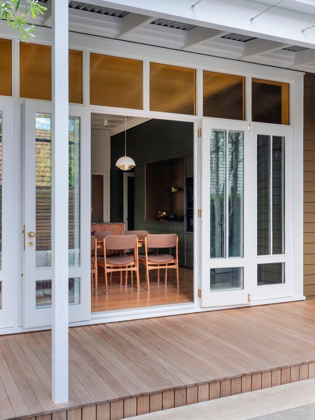 A bright covered timber deck opens through white-painted glazed folding doors into a dining and living area. The interior features a timber dining table and chairs, dark green built-in cabinetry, warm timber flooring, and a pendant light. The exterior door surround includes amber textured glass transom panels, while the deck has timber flooring and a simple white post-and-beam structure.