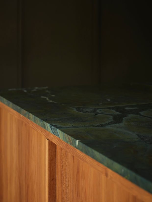 Close-up detail of a dark, heavily veined natural stone benchtop with a thick edge profile, set above warm-toned timber cabinetry with vertical recessed detailing. The subdued lighting and dark wall create a moody, refined interior finish.