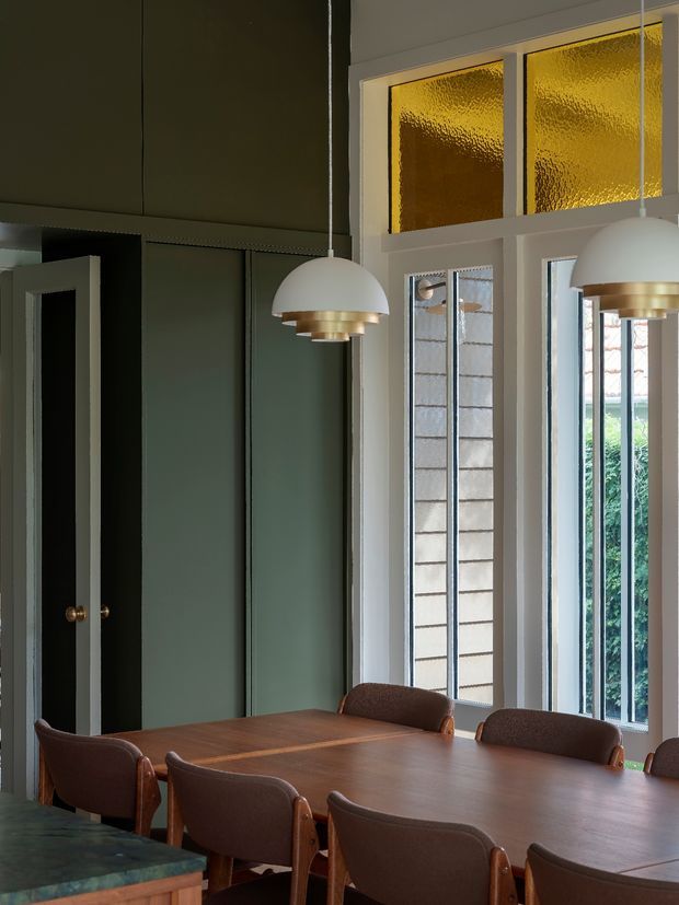 A refined dining room with a long dark timber dining table surrounded by brown upholstered chairs. The room features deep green built-in wall panelling and cabinetry, tall white-framed windows and doors, amber textured clerestory glazing, and white dome pendant lights with brass details. The overall atmosphere is moody, warm, and sophisticated.