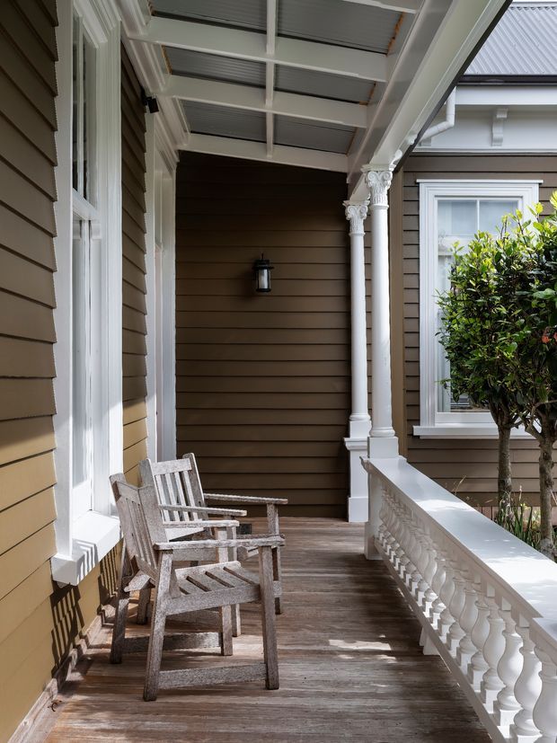 A covered traditional front veranda with weatherboard walls, white decorative columns and balustrade, timber decking, tall white-trimmed windows, and two wooden outdoor chairs. The space has a refined heritage character with warm brown cladding and abundant natural light.