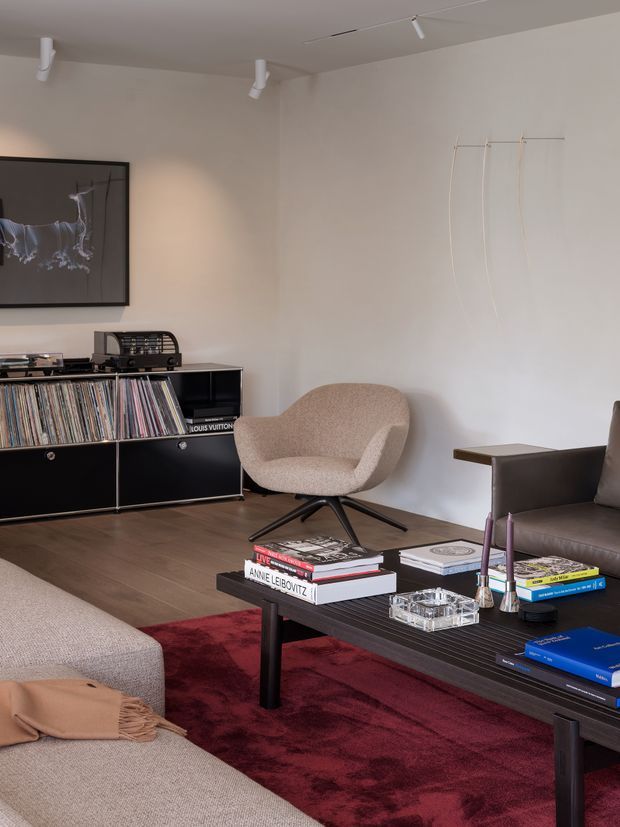 A refined contemporary living room with warm neutral walls, dark timber flooring, and a deep burgundy rug. The room features a black record cabinet filled with vinyl records, a sculptural upholstered lounge chair, a dark leather sofa, and a low black coffee table styled with art books and decorative objects. Track lighting and understated wall art complete the curated, music-oriented interior.