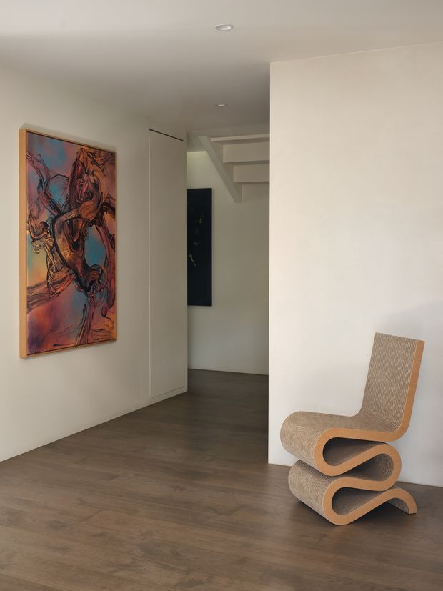 A minimalist interior hallway or entry space with warm dark timber flooring, smooth off-white walls, recessed ceiling lights, and a sculptural woven wooden chair. A colourful framed artwork hangs on the left wall, while an opening reveals a staircase and darker interior beyond.