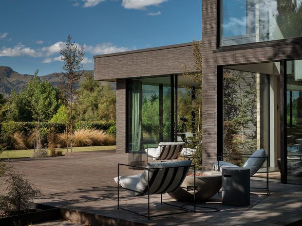 Contemporary flat-roofed residence with dark textured masonry cladding and extensive floor-to-ceiling glazing, opening onto a spacious paved outdoor terrace. Minimal outdoor lounge furniture sits beside landscaped planting, with mountainous terrain visible beyond the property.
