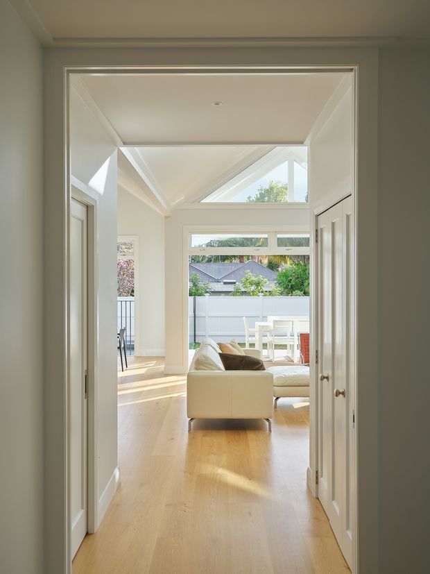 A bright interior view through a wide doorway into an open-plan living area. The space features pale timber flooring, white walls and trim, a cream sectional sofa, large windows, and a glazed clerestory-style roof section that fills the room with natural light. A dining or outdoor seating area and landscaped suburban surroundings are visible beyond the glazing.
