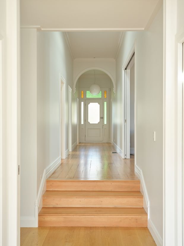 Bright, refined residential hallway and entryway viewed from the lower level, with a short flight of warm timber stairs leading to a long corridor. The corridor has polished timber flooring, pale painted walls, white decorative skirting and architraves, an arched opening, high detailed ceilings, and a traditional front door with narrow coloured glass sidelights and transom. Multiple doorways open from the hall.