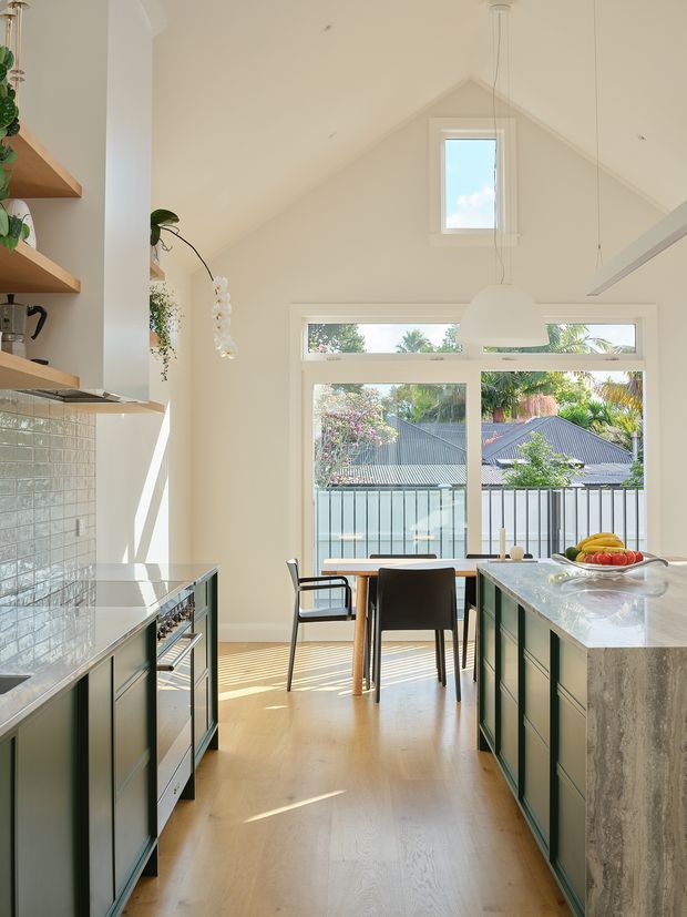 Bright contemporary kitchen with a tall pitched ceiling and prominent clerestory-style glazing. Pale timber flooring, dark green fixed cabinetry, light stone benchtops, and a glossy light subway-tile splashback frame a central island and dining area. Large windows provide views to neighbouring rooftops and lush vegetation, while pendant lighting and open timber shelving add warmth.