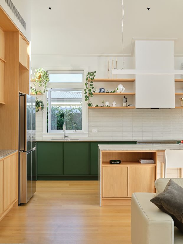 Bright contemporary kitchen with a warm timber interior, deep green lower cabinetry, white tiled splashback, open timber shelving, stainless-steel appliances, and a large white rangehood. A living area with a cream sofa is partially visible in the foreground, while a window brings in abundant natural light.