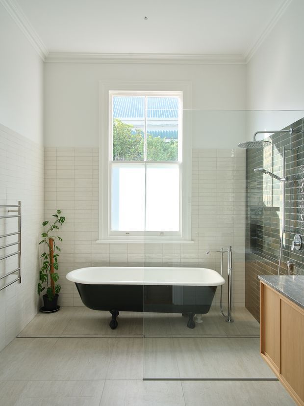Bright, spacious bathroom with a freestanding black-and-white clawfoot bathtub centered beneath a sash window. The room features pale large-format floor tiles, white horizontal wall tiles, white decorative ceiling cornices, a frameless glass shower enclosure with chrome fixtures, a timber vanity with a dark stone benchtop, and a heated towel rail. A small potted plant adds greenery.
