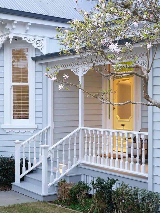 Exterior view of a finely detailed traditional timber-clad house entry. The pale blue weatherboard facade features ornate white decorative fretwork, corbels, trim, turned porch balusters and a small covered verandah reached by steps. A tall narrow window with timber blinds sits beside a warm yellow front door, with flowering magnolia branches in the foreground.