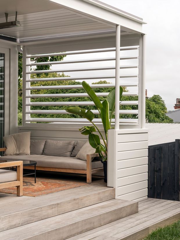 A bright, covered outdoor living area with a timber-framed sofa, cushions, coffee table and patterned rug. The space is enclosed by white horizontal louvres and privacy screening, with a tall tropical plant in a built-in planter. Pale timber decking and broad steps lead down to a fenced garden area.