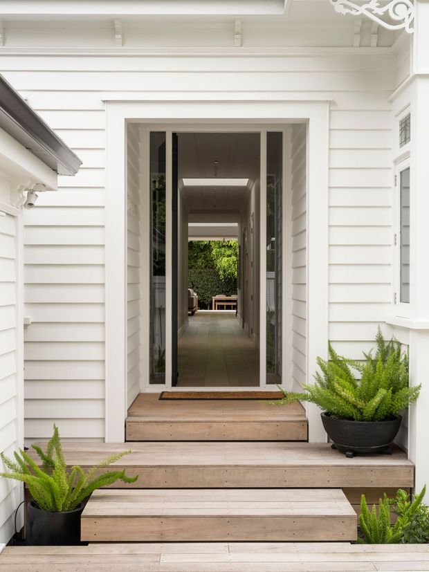 Bright exterior entry to a white weatherboard-clad home, featuring a glazed front door with sidelights, timber steps and deck, and potted ferns. The doorway opens to a long interior corridor leading toward a leafy courtyard or garden beyond.