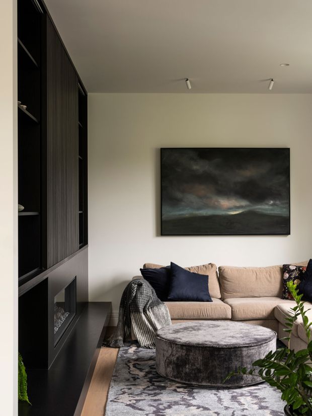 A refined contemporary living room with a beige sectional sofa, navy and patterned cushions, a large round upholstered ottoman, and a monochrome patterned rug. A dark built-in media wall with open shelving and a fireplace frames the left side, while a large moody landscape artwork hangs on the pale wall. Track and recessed lighting provide soft illumination, and indoor greenery adds a natural accent.