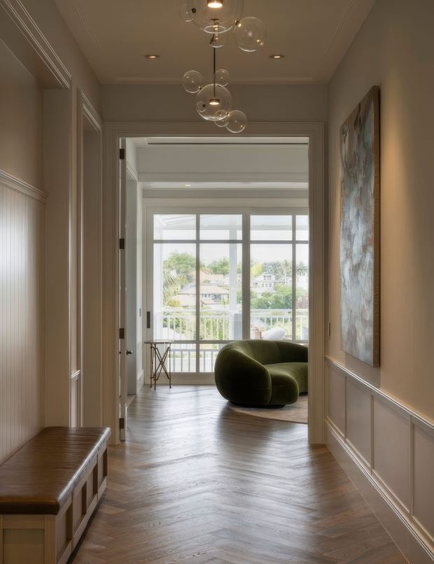 A refined residential hallway with warm taupe walls, decorative wall panelling and trim, and a dark timber herringbone floor. A built-in timber bench lines the left wall, while a sculptural glass pendant hangs overhead. Through the open doorway is a bright living area with floor-to-ceiling glazing, a curved olive-green sofa, and a balcony overlooking leafy suburban surroundings. An abstract artwork provides a restrained feature on the right wall.