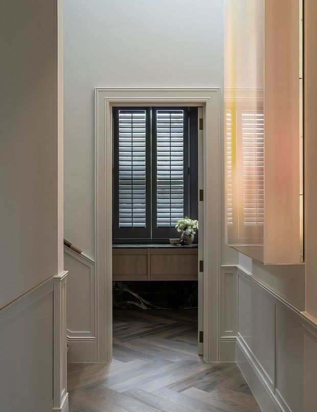 A refined residential hallway with white painted wall panelling and moulded trim leads through an open doorway to a compact room featuring a dark-framed window with plantation shutters, a timber vanity or built-in cabinet, and herringbone timber flooring. Warm light and layered classical detailing create an elegant transitional atmosphere.