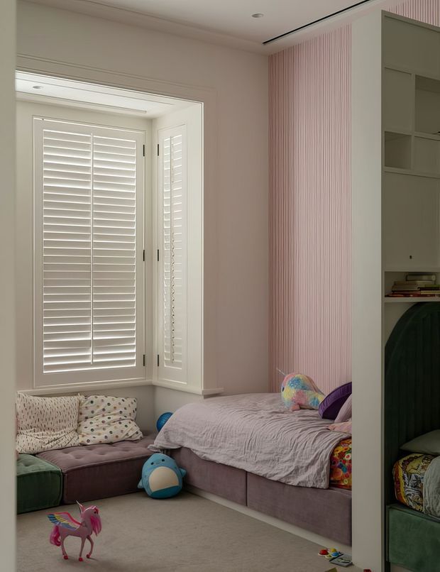 A softly lit children's bedroom with a built-in window seat beneath white plantation shutters, a mauve upholstered bed, plush seating, colourful cushions and toys. The room features pale pink vertical ribbed wall detailing and integrated built-in shelving and wardrobe storage on the right.