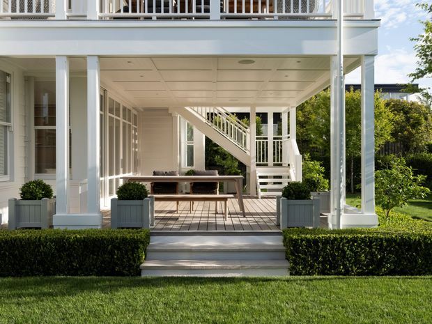 A bright, formal outdoor terrace beneath a raised white veranda, with square columns, white balustrades, a covered ceiling, timber decking, built-in seating, a dining table, potted shrubs, clipped hedges, and steps leading to a manicured lawn. A staircase rises through the veranda to the upper level, reinforcing the multi-storey residential architecture.