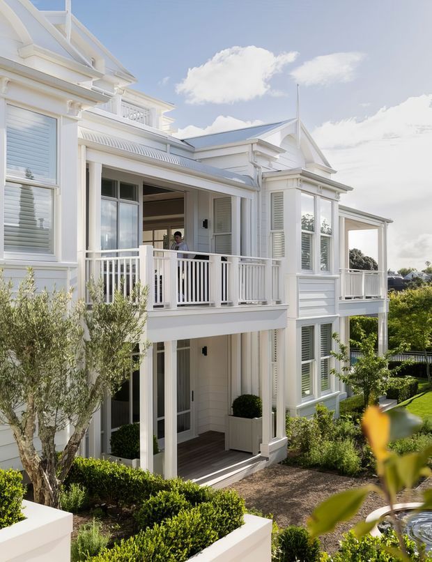Bright exterior view of a substantial two-storey white coastal-style residence with ornate classical detailing, pitched roof elements, dormer-like gables, covered balconies, white balustrades, louvred shutters, and extensive white-painted trim. The house is surrounded by manicured hedges, raised garden beds, gravel paths, and established landscaping. A small portion of a water feature is visible in the foreground.