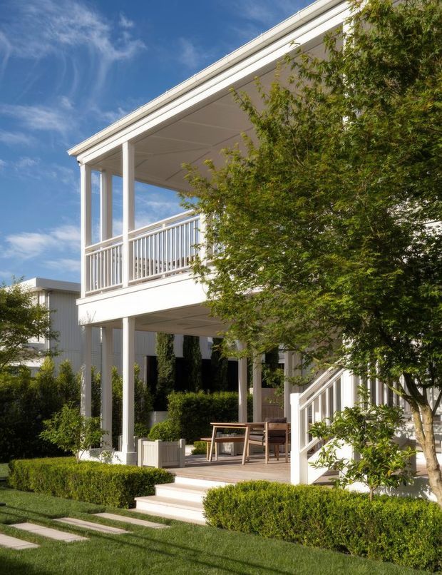 Exterior view of a bright, elevated two-storey residential building with a broad covered upper balcony, white vertical columns and railings, open ground-level patio, and landscaped garden with clipped hedges and mature trees. The clean white facade and generous outdoor living areas are prominent.