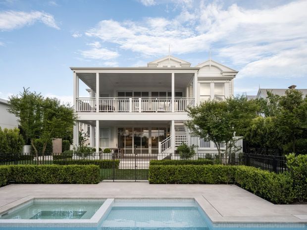 Front exterior view of a large white, two-storey residence with a covered upper balcony, classical gabled detailing, verandah-style columns and railings, manicured hedges, a gated garden, and a foreground swimming pool with spa.