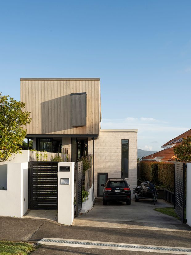 Exterior street view of a contemporary multi-level urban residence with a cantilevered upper storey clad in vertical timber battens, pale brick lower walls, dark-framed glazing, and a narrow driveway leading to a garage or parking area. The entry includes a black slatted pedestrian gate, rendered boundary walls, mailbox, and house number. Neighbouring homes and distant hills are visible in the background.