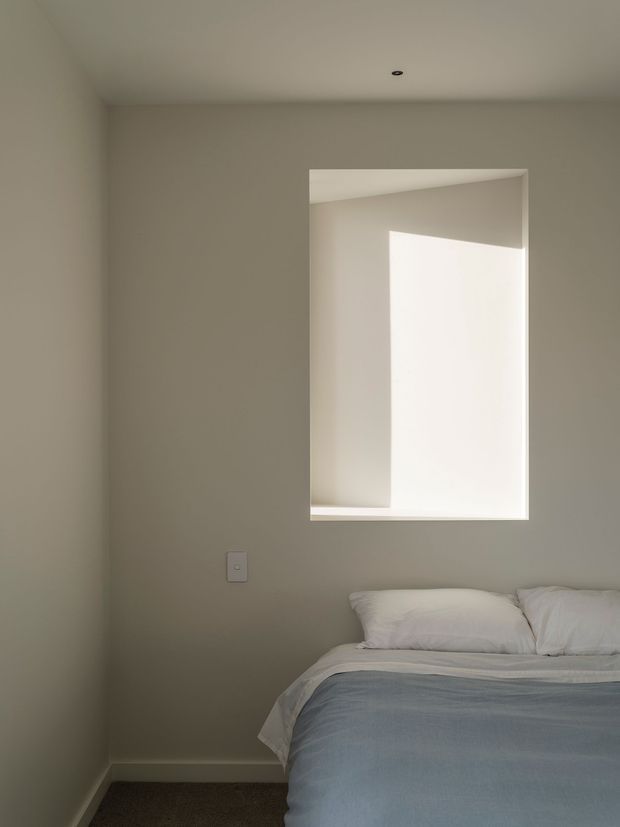 A minimalist bedroom corner with a neatly made bed partially visible in the lower right. The room has warm light-grey walls, a plain white ceiling with a small recessed downlight, white skirting boards, and a deep-set rectangular window recess that admits bright natural light. Bedding is white with a muted blue-grey cover, creating a calm and understated atmosphere.