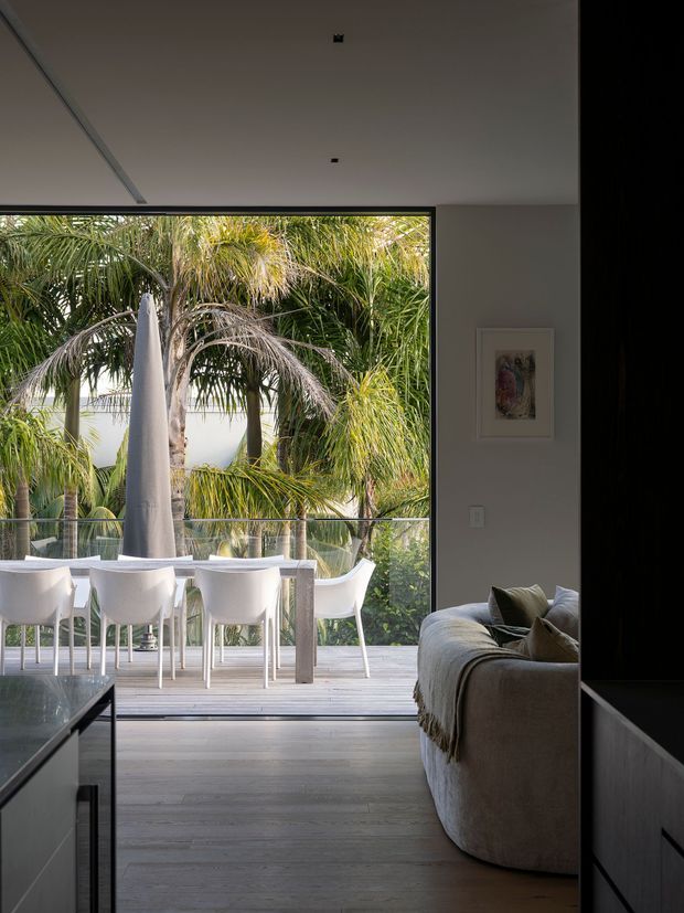 A contemporary open-plan living and dining space opening onto a timber deck. The image is framed from the living area, with a large grey upholstered sofa in the foreground and a white dining table with multiple white chairs beyond. Full-height black-framed glazing connects the room to a lush tropical garden and poolside setting. The interior has a restrained neutral palette, pale timber flooring, a smooth white ceiling with recessed lights, and minimal wall art.