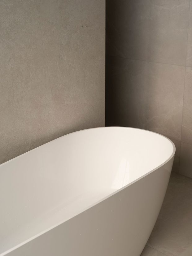 Close-up of a minimalist bathroom featuring a freestanding white oval bathtub against large-format warm grey-beige tiled walls and floor. The restrained palette, smooth surfaces, and soft natural-looking light create a calm, refined atmosphere.