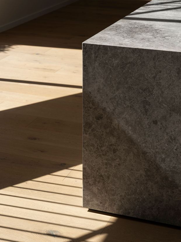 Close-up architectural detail of a dark grey, heavily veined stone island or benchtop with a thick waterfall-style end panel. The surface is lit by strong sunlight and window shadows, set against a pale timber floor. No wider room context or cabinetry is visible.