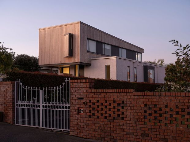Exterior view of a contemporary two-storey house at dusk, featuring a box-like upper volume clad in vertical timber, pale brick lower-level walls, broad dark-framed windows, and a projecting flat roof. A patterned brick boundary wall and ornate metal driveway gate occupy the foreground.