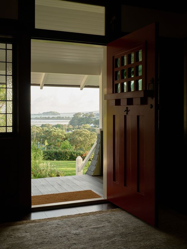 A dimly lit entryway looking through an open, dark-stained timber front door with small divided glass panes. The covered porch has a white-lined ceiling and grey decking, opening onto a landscaped garden and a broad coastal or waterfront view beyond. A sidelight window with divided panes is visible beside the door, while a textured rug and doormat sit inside the threshold.
