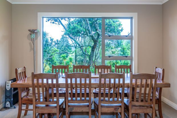 A traditional dining room centered on a long solid-wood dining table surrounded by numerous wooden slat-back chairs. The room has warm taupe painted walls, white crown moulding and skirting, and a large white-trimmed window overlooking dense greenery. A slim floor lamp and portable heater sit along the side wall. The space is bright, warm, and family-oriented, with a strong connection to the garden outside.