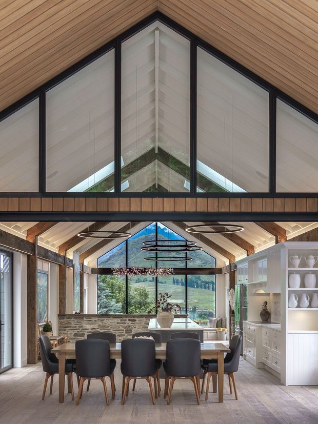 A refined dining room beneath a dramatic pitched, gabled ceiling with extensive black-framed glazing overlooking a green landscape. The room features warm timber ceiling lining and structural beams, a long dining table with upholstered dark chairs, rustic stone wall cladding, pale timber flooring, sculptural ring pendant lights, and a large white built-in display cabinet. A prominent upper-level gable window and exposed roof structure create a strong architectural focal point.