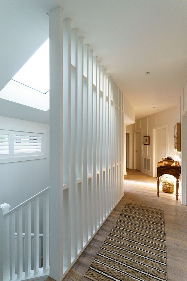 Bright upper-level hallway or landing with pale timber flooring, white walls, and a distinctive full-height white vertical-slat balustrade screening an open stairwell. Natural light enters through a skylight and shuttered window, while a striped runner, console table, framed artwork, and warm decorative lighting add detail along the corridor.