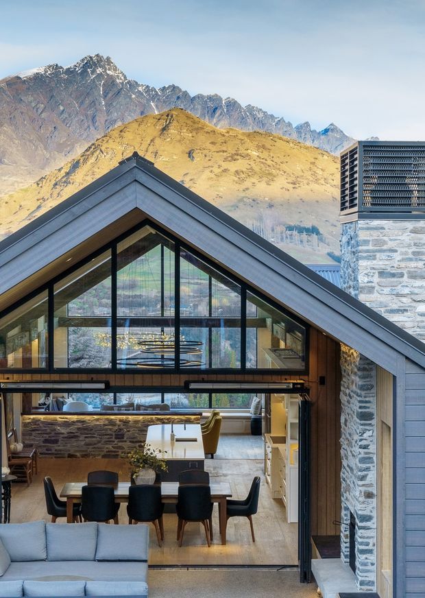 A dramatic alpine residence with a steep gabled roof and extensive black-framed glazing. The open-plan interior features a dining area, kitchen, stone island, timber cabinetry, a lounge, and a mezzanine-like upper level. Rough natural-stone walls, timber lining, polished light flooring, and sculptural pendant lighting create a warm contemporary mountain retreat, with rugged snow-dusted peaks visible beyond.